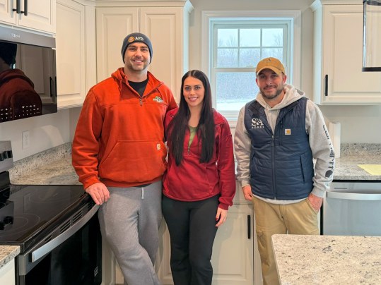 Three people standing in a bright white kitchen with granite countertops, black appliances, and large window behind them
